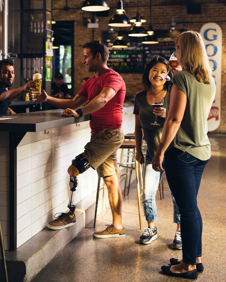A group of friends talking and drinking together at a bar, one standing with a prosthetic leg