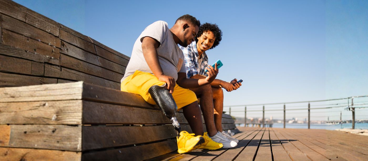 Two people sitting on a boardwalk in the sun, one with a prosthetic leg