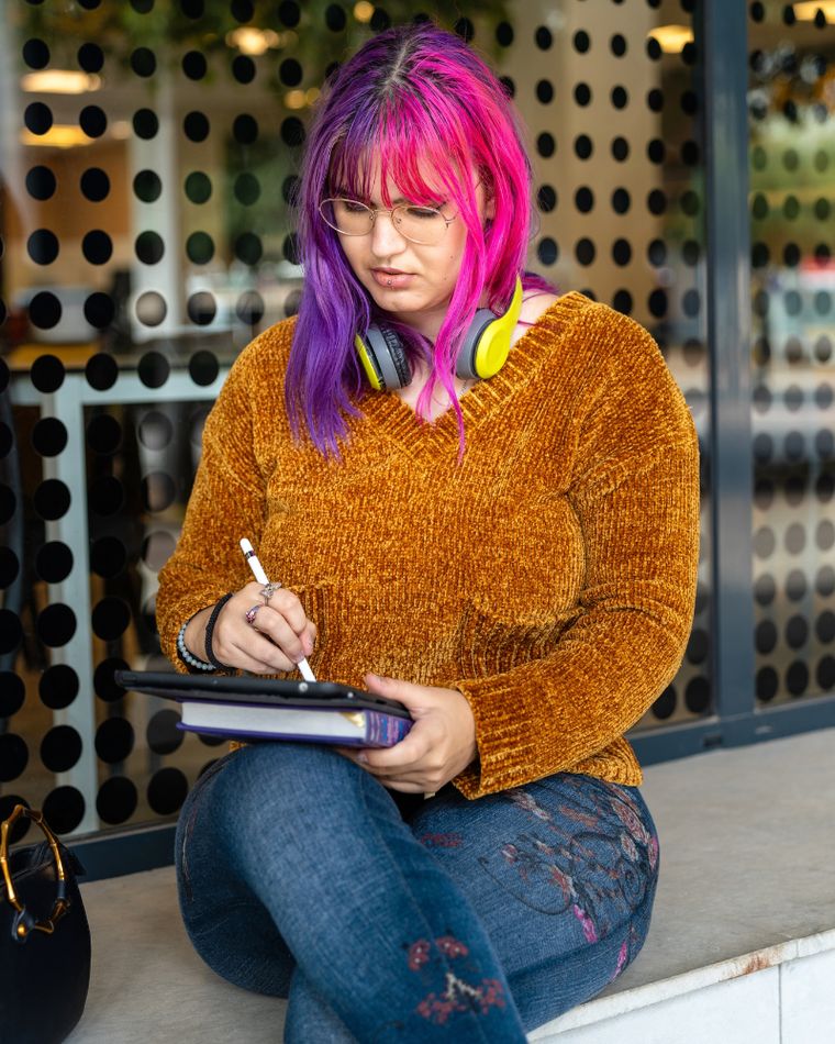 A person with brightly coloured hair sitting and writing, headphones around their neck