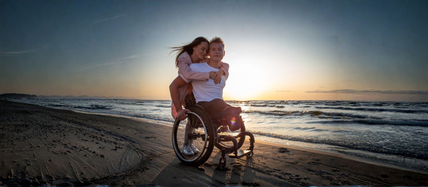 A couple together on a beach at sunset, one using a wheelchair