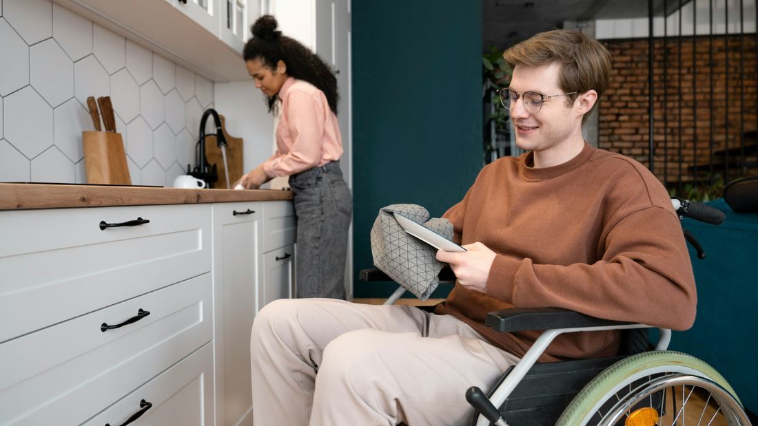 A man in a kitchen at home, wheelchair alongside, reading his phone