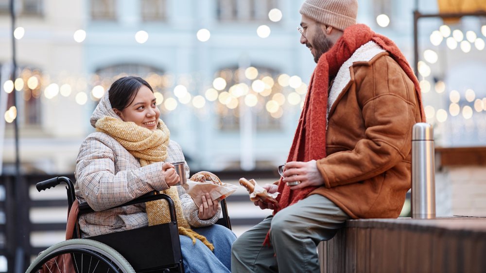A couple talking and sharing a hot drink at an outdoor market in the evening, one of them using a wheelchair